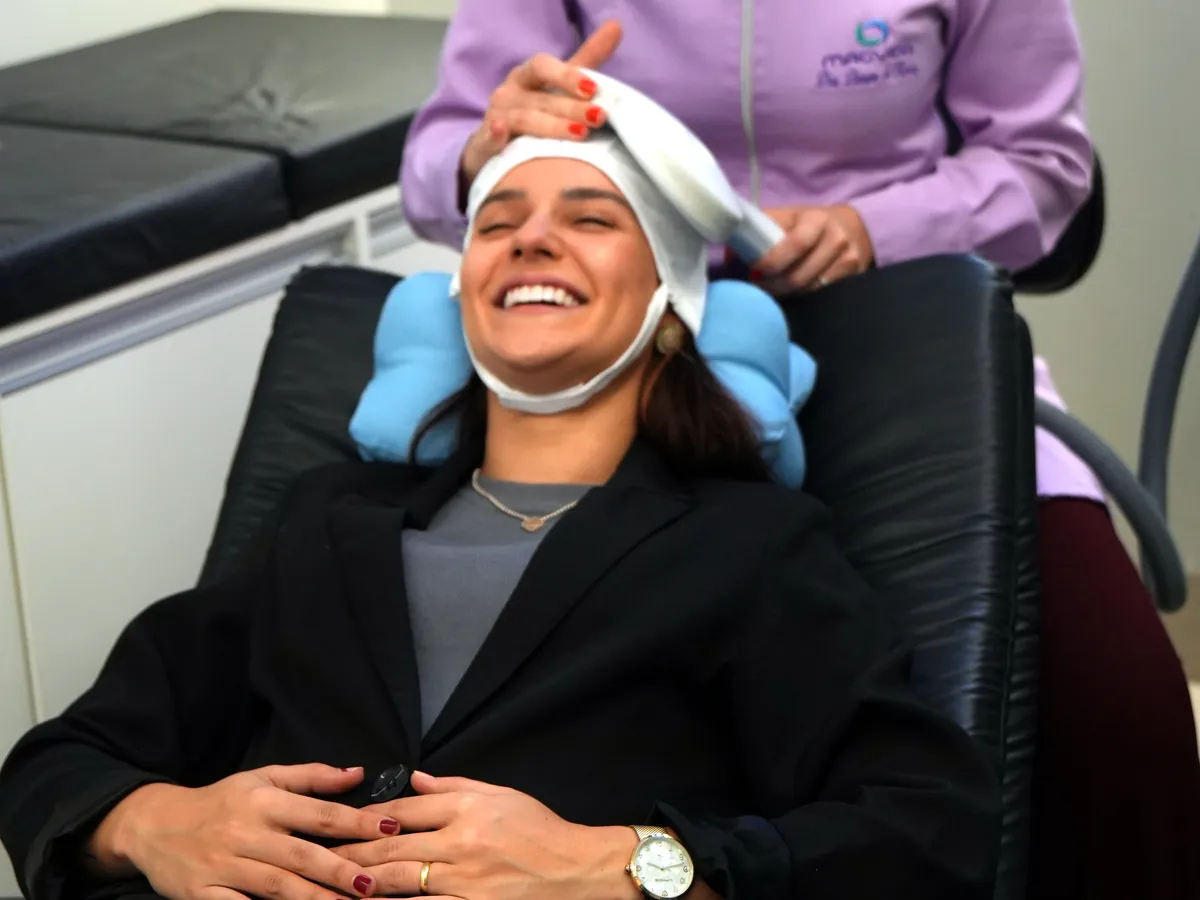 Paciente sorrindo na poltrona durante a sessão, com a fisioterapeuta ao lado